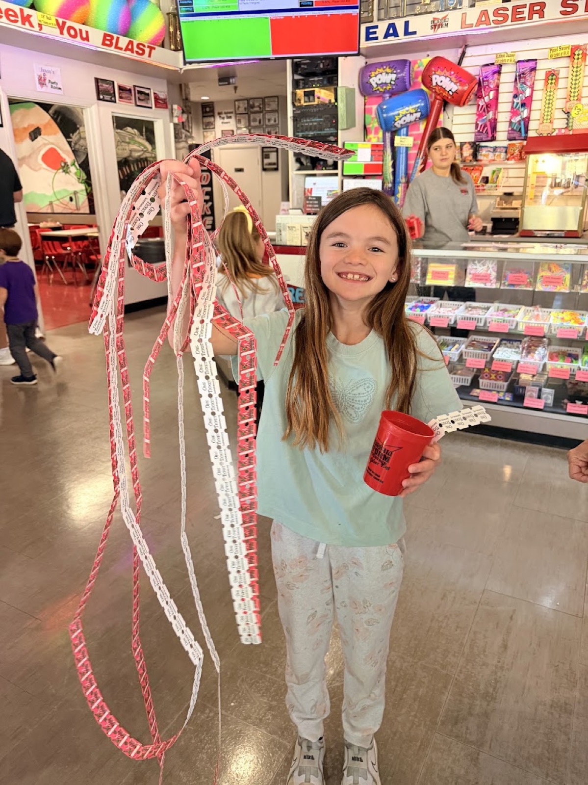 A grinning kid holding up long, tangled strands of prize tickets in one hand and a red cup of tokens in the other
