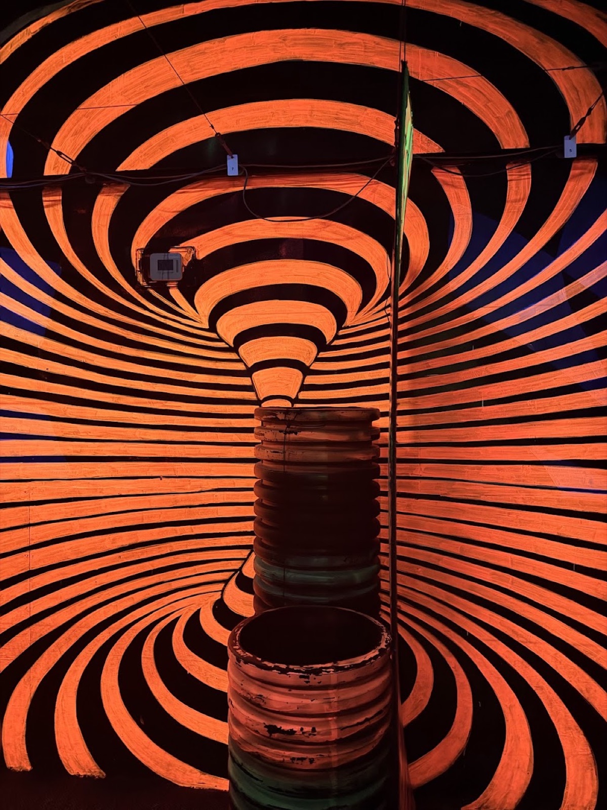 A floor-to-ceiling orange-and-black funnel painting that makes a stack of barrels look like they're being sucked into a wormhole